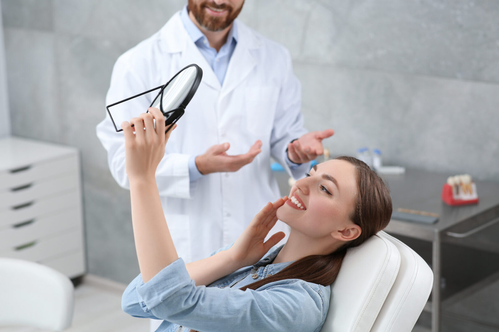 Costa Mesa Family Dentistry with Dr Almodovar | A woman sitting in a dental chair smiles while holding a mirror. A dentist in a white coat gestures with his hands. The room has dental equipment.
